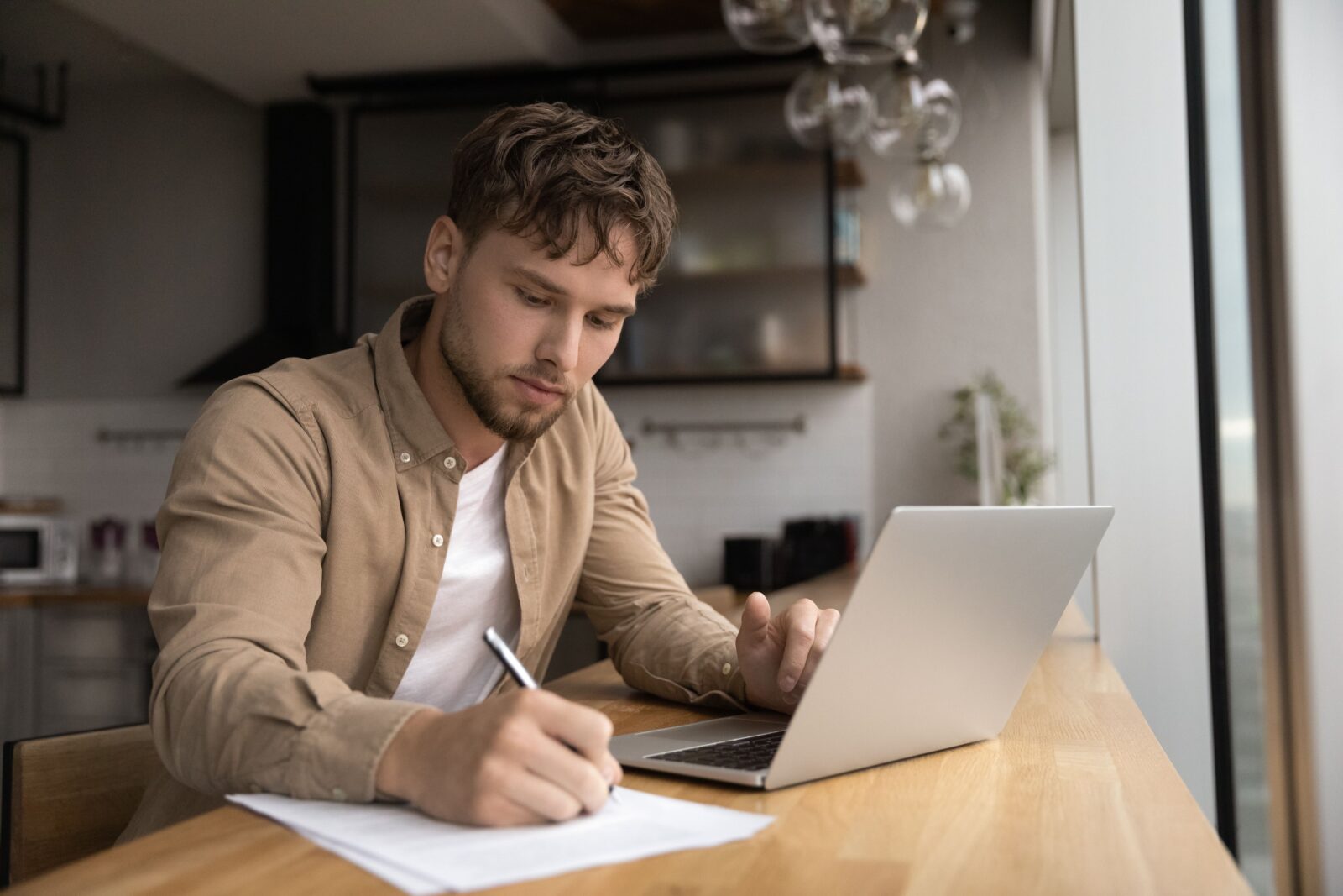 Joven estudiante prepara un ensayo de investigación en una cocina moderna, navega por internet, usa una laptop y escribe información en papel para un informe a mano. Hombre independiente enfocado que trabaja desde casa, elaborando una lista de datos desde la pantalla de su computadora